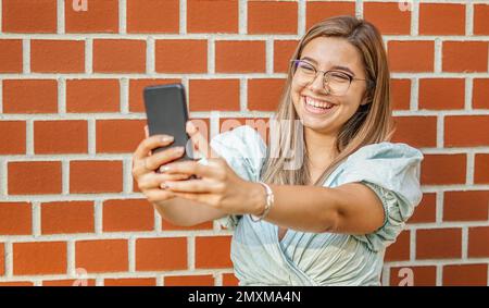 Jeune fille prenant un selfie - souriant à l'appareil photo - heureux étudiant debout près du mur et en utilisant son smartphone Banque D'Images