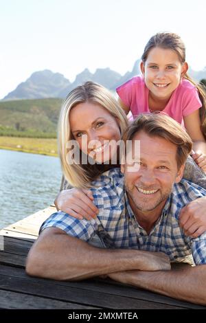 Des souvenirs qui durent toute une vie. Portrait d'une famille heureuse de trois personnes assises ensemble au bord d'un lac. Banque D'Images