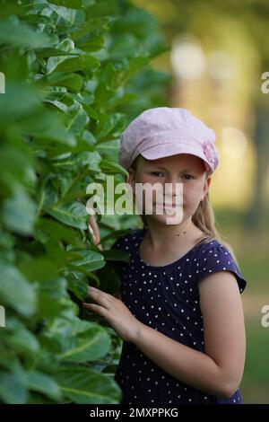 Mignon caucasien petite fille dans un chapeau rose debout près de la fleur de la buisson de l'hortensia bleu. Mise au point sélective Banque D'Images