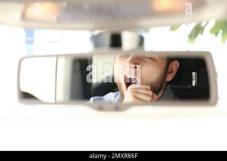 Homme fatigué qui bâille pendant la conduite, réfléchissant dans le rétroviseur intérieur Banque D'Images