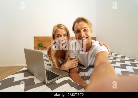 Un jeune couple heureux qui s'amuse à prendre le selfie tout en utilisant un ordinateur portable au lit - concept de relation d'amour et de technologie Banque D'Images