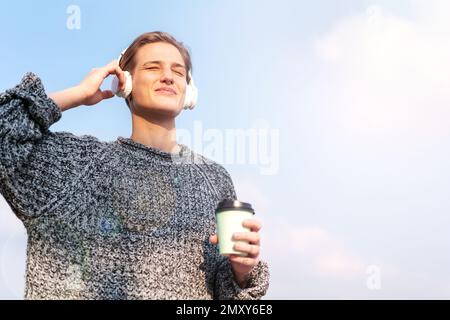 Femme debout à l'extérieur contre le ciel bleu tenant une tasse à café jetable dans la main écoutant de la musique à l'aide d'un casque sans fil et souriant. Banque D'Images