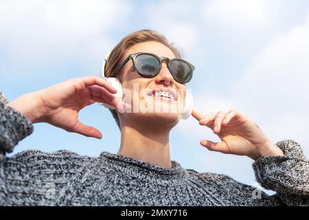 Portrait vue à angle bas jeune cheveux courts femme en verre de soleil écouter de la musique à l'aide de grands écouteurs et sourire. Banque D'Images