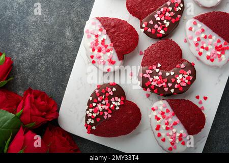 Des biscuits au velours rouge ou au brownie sur le cœur en forme de glaçage au chocolat avec des roses rouges sur fond noir. Idée de dessert pour la Saint-Valentin, les mères ou les femmes Banque D'Images