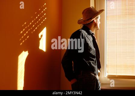 Un homme dans un chapeau et une cravate regarde à travers un aveugle à la fenêtre dans la lumière du soir Banque D'Images