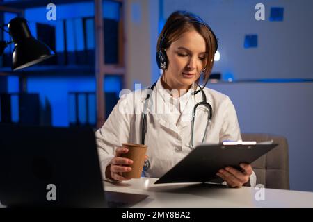 Jeune médecin fatigué travaillant de nuit au bureau avec des papiers, thérapeute avec casque tenant un appel vidéo avec le patient Banque D'Images