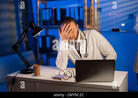 Médecin surpassé fatigué avec migraine de travail de nuit en utilisant un ordinateur portable tout en étant assis à une table Banque D'Images