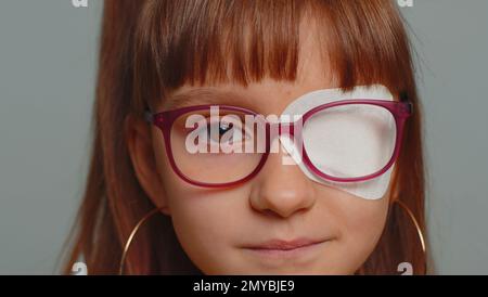 Gros plan portrait malade préteen fille ayant le pansement protecteur patch scratch sur un œil. Blessure, traumatisme, aveugle. Enfant enfant portant des lunettes sur fond gris. Traitement médical de la vue Banque D'Images