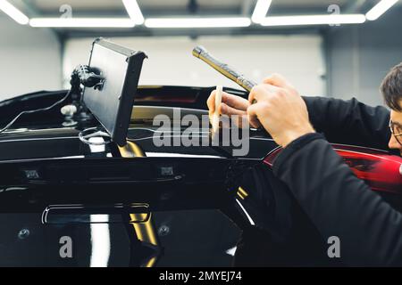 Homme qui retire une bosselure sur le couvercle de coffre noir. Réparation de rente sans peinture professionnelle. Gros plan en intérieur. Atelier de mécanique et de détail de voiture. Photo de haute qualité Banque D'Images