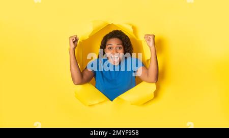 Une femme noire surjoyeuse qui applaudit les poings serrés, célébrant la victoire, posant dans un trou déchiré de fond jaune Banque D'Images