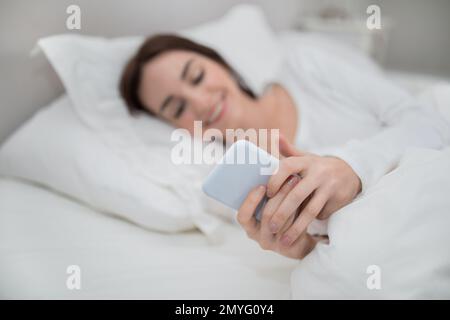 Smartphone dans les mains de jeune femme souriante, intérieur de chambre Banque D'Images
