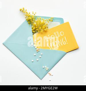 Légende Hello Spring sur carton jaune, fleurs mimosa fraîches dans enveloppe bleu menthe sur fond blanc. Flat lay, vue de dessus, composition centrale Banque D'Images