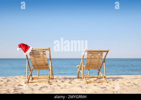 Chaises longues et chapeau de Père Noël sur la plage, espace pour le texte. Vacances de Noël Banque D'Images