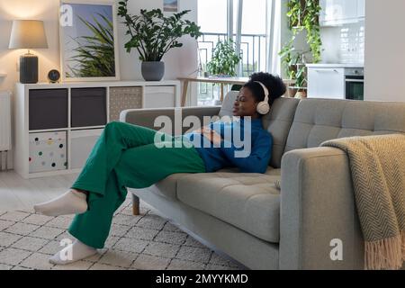 Bonne femme afro-américaine décontractée dans un casque qui ferme les yeux en écoutant de la musique assis sur un canapé Banque D'Images