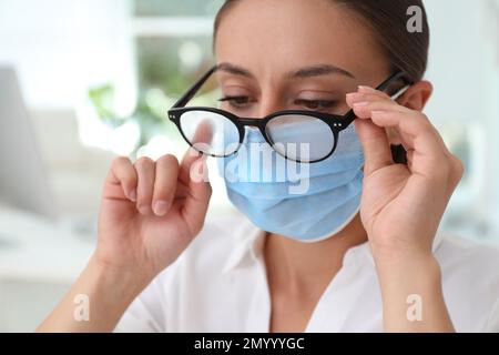 Femme essuyant des lunettes brumeuses causées par le port d'un masque médical à l'intérieur, gros plan Banque D'Images