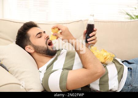 Un jeune homme paresseux avec des frites et des boissons sur le canapé à la maison Banque D'Images