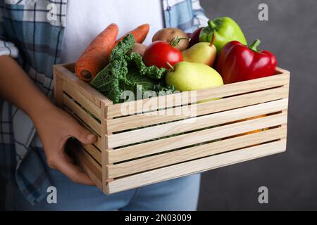 Fermier tenant une caisse en bois remplie de légumes frais et de fruits sur fond gris, gros plan Banque D'Images