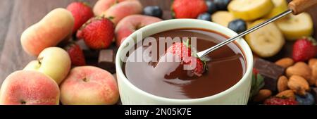 Fourchette à fondue avec fraise dans un bol de chocolat fondu entouré d'autres fruits sur une table en bois Banque D'Images