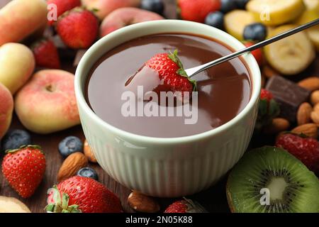 Fourchette à fondue avec fraise dans un bol de chocolat fondu entouré d'autres fruits sur une table en bois Banque D'Images