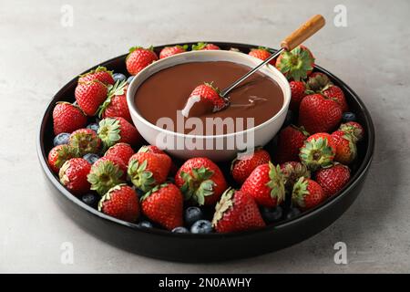 Fourchette à fondue avec fraise dans un bol de chocolat fondu entouré de différentes baies sur table grise Banque D'Images