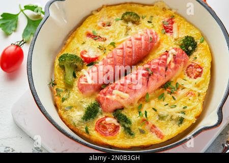 Omelette ou frittata avec deux saucisses, fromage fondu, tomates cerises, brocoli et persil sur poêle rouge ou poêle en fer sur fond blanc en béton Banque D'Images