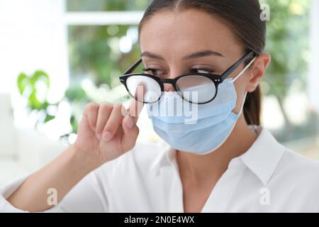 Femme essuyant des lunettes brumeuses causées par le port d'un masque médical à l'intérieur, gros plan Banque D'Images