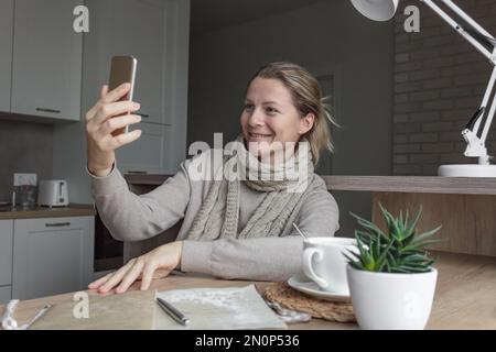 Une femme dans un chandail et une écharpe tricotée dans la cuisine s'assoit à une table en bois et parle sur un appel vidéo au téléphone. Banque D'Images