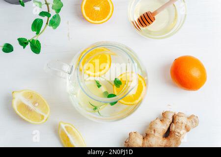 Théière en verre avec thé à l'orange, au citron, au gingembre et à la menthe avec ingrédients sur une table en bois blanc. Vue de dessus, plan d'agencement. Banque D'Images