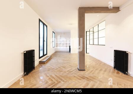 Chambre vide d'un appartement de style loft avec murs peints en blanc, radiateurs en fonte noire, fenêtres en fer avec cloisons en verre et pilier en béton brut Banque D'Images