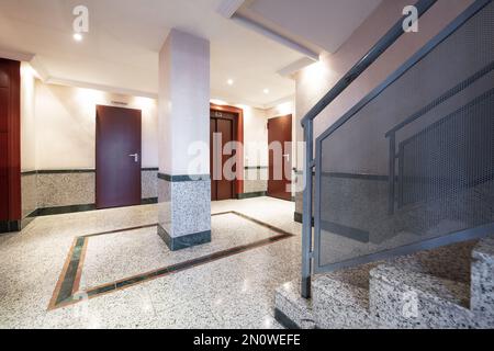 Grand portail d'entrée vers un bâtiment résidentiel avec des escaliers en granit de différents tons, des murs en demi-carreaux et un ascenseur avec des portes rouges Banque D'Images