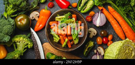 Salade de brocolis de carottes vapeur dans un bol et assortiment de légumes frais coupés sur table en béton rustique, planche à découper en bois, couteau de cuisine d'en haut, légumes Banque D'Images