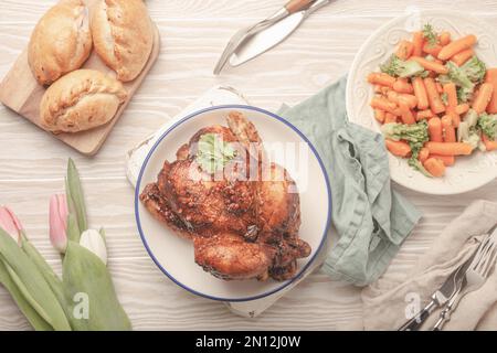 Repas de Pâques sur table rustique blanche : poulet rôti et légumes, petits pains et fleurs de printemps tulipes vue de dessus plat, repas de Pâques en famille avec fe Banque D'Images