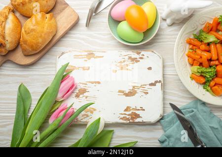 Tableau blanc en bois vide et nourriture de Pâques sur table de cuisine: Œufs pastel, poulet rôti, légumes, petits pains, tulipes de printemps vue du dessus, EAS Banque D'Images