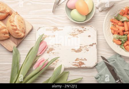 Tableau blanc en bois vide et nourriture de Pâques sur table de cuisine: Œufs pastel, poulet rôti, légumes, petits pains, tulipes de printemps vue du dessus, EAS Banque D'Images