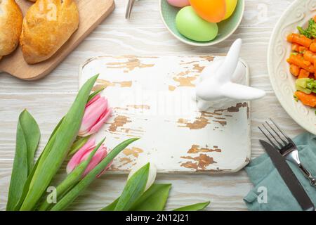 Tableau blanc en bois vide et nourriture de Pâques sur table de cuisine: Œufs pastel, poulet rôti, légumes, petits pains, tulipes de printemps vue du dessus, EAS Banque D'Images