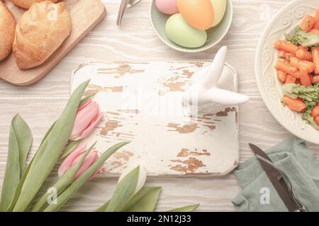 Tableau blanc en bois vide et nourriture de Pâques sur table de cuisine: Œufs pastel, poulet rôti, légumes, petits pains, tulipes de printemps vue du dessus, EAS Banque D'Images