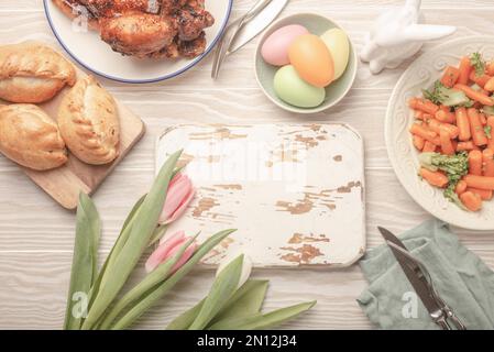Tableau blanc en bois vide et nourriture de Pâques sur table de cuisine: Œufs pastel, poulet rôti, légumes, petits pains, tulipes de printemps vue du dessus, EAS Banque D'Images