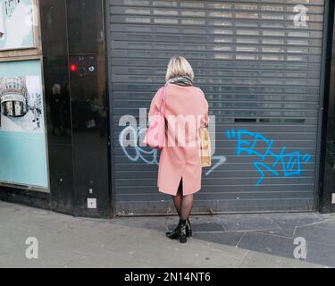 Femme dans un manteau de pêche regarde le graffiti vaporisé sur un magasin une obturateur dans une rue de Londres Angleterre Banque D'Images