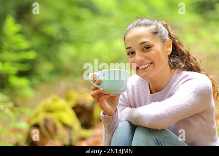 Femme heureuse assise dans la nature tenant une tasse de café posant regardant l'appareil photo Banque D'Images