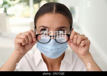 Femme essuyant des lunettes brumeuses causées par le port d'un masque médical à l'intérieur, gros plan Banque D'Images