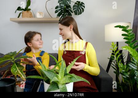 Belle jeune mère et heureuse petite fille adorable enfant essuyant la poussière des feuilles de la maison, prenant soin de la plante assis sur le canapé à la maison. Accueil ga Banque D'Images