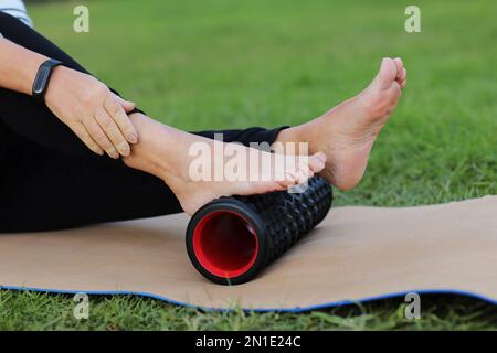 Journée mondiale de la santé. Une femme méconnaissable se réchauffe et travaille le pied des muscles du corps avec un rouleau de mousse de massage sportif à l'extérieur sur le g vert Banque D'Images