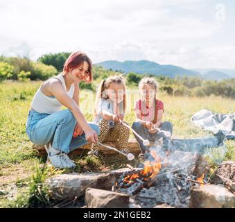 Trois sœurs sourient en rôtisant un guimauve de bonbons sur les bâtons au-dessus de la flamme du feu de camp. Joyeux pique-nique en famille ou en plein air Banque D'Images