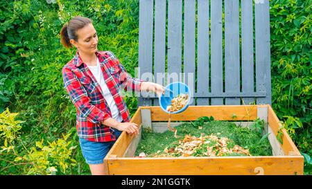 Bac à compost en bois à fabriquer soi-même. Une femme jette des déchets alimentaires dans un bac à compost pour préparer un sol fertile pour un jardin biologique. Déchets de cuisine, pelouse tondue Banque D'Images