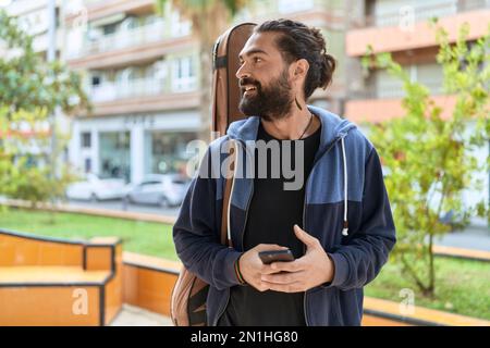Jeune homme hispanique musicien utilisant un smartphone tenant une caisse de guitare au parc Banque D'Images