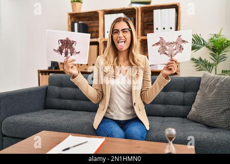 Jeune femme blonde thérapeute tenant le test rorschach collant la langue dehors heureux avec l'expression drôle. Banque D'Images