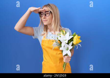 Jeune femme caucasienne portant un tablier fleuriste tenant des fleurs très heureux et souriant regardant loin avec la main sur la tête. concept de recherche. Banque D'Images