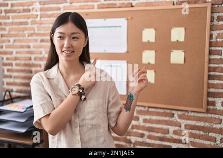 Jeune femme chinoise travaillant au bureau faisant la présentation souriant et regardant la caméra pointant avec deux mains et des doigts sur le côté. Banque D'Images