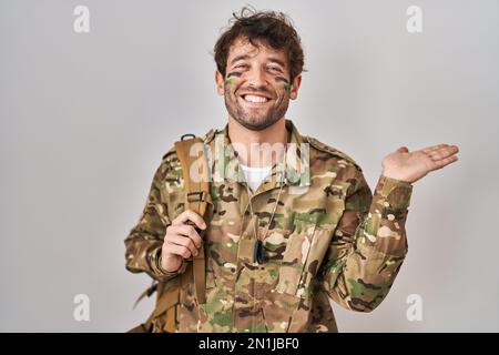 Jeune homme hispanique portant un uniforme militaire de camouflage souriant gai présenter et pointer avec la paume de la main regardant la caméra. Banque D'Images
