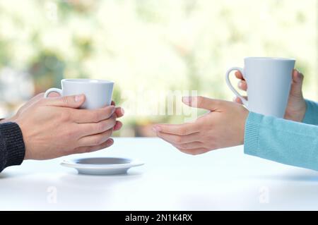 Profil de rétroéclairage des mains de deux amis ou d'un couple parlant tenant des tasses de café assis dans une table à la maison avec une fenêtre en arrière-plan Banque D'Images
